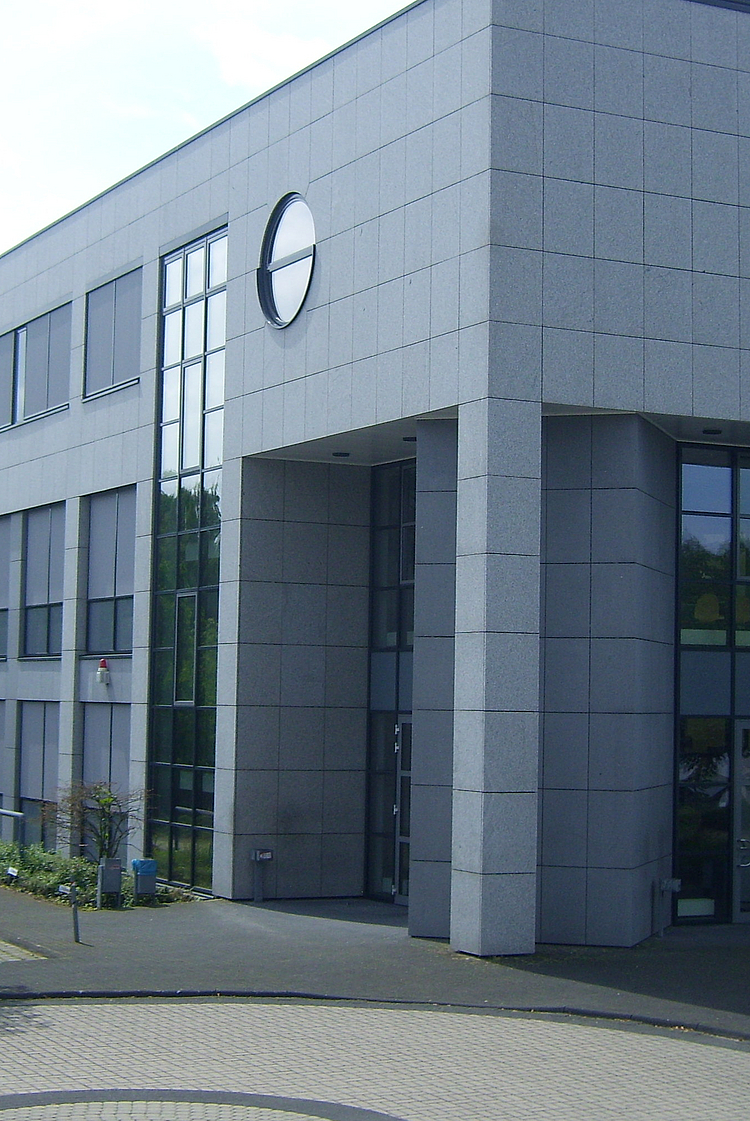 Front 001 Ein modernes Bürogebäude mit einer glatten Steinfassade und großen Fenstern. Vor dem Gebäude befinden sich einige Bäume und ein Parkplatz. Es gibt eine auffällige runde Fensterform auf der Fassade. Das Bild ist an einem sonnigen Tag aufgenommen.